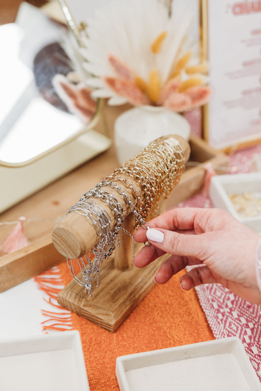 Person holding a silver bracelet next to a wooden jewelry stand with more bracelets on a decorative table.