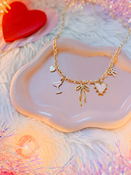 Gold necklace with heart, bow, and martini glass charms on a pink dish with a heart-shaped object in the background.