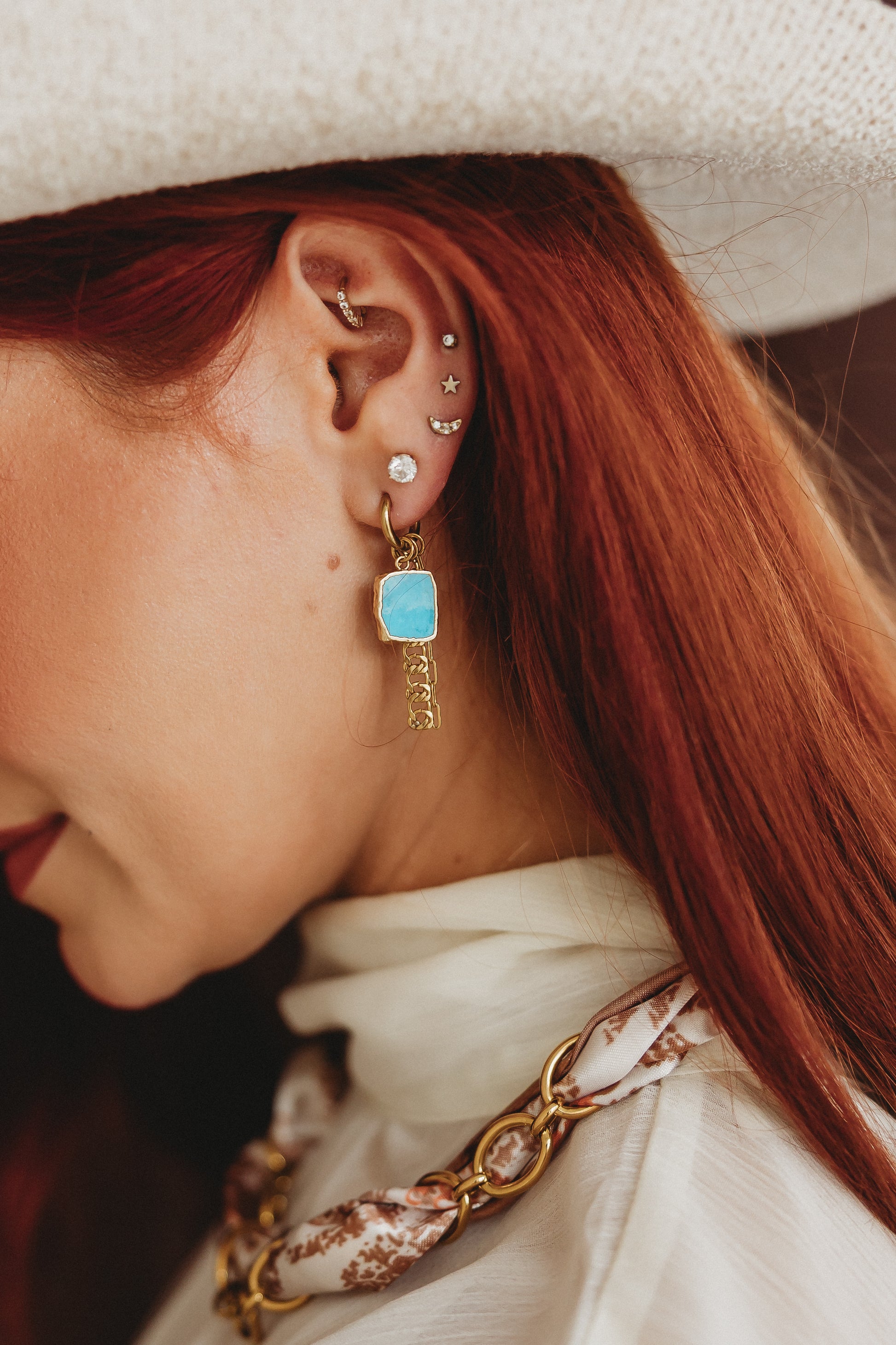 A close-up side profile of a woman with red hair wearing a white wide-brim hat, white top, and a gold chain necklace, highlighting a dangling earring with a light blue rectangular stone and gold chain dangle.
