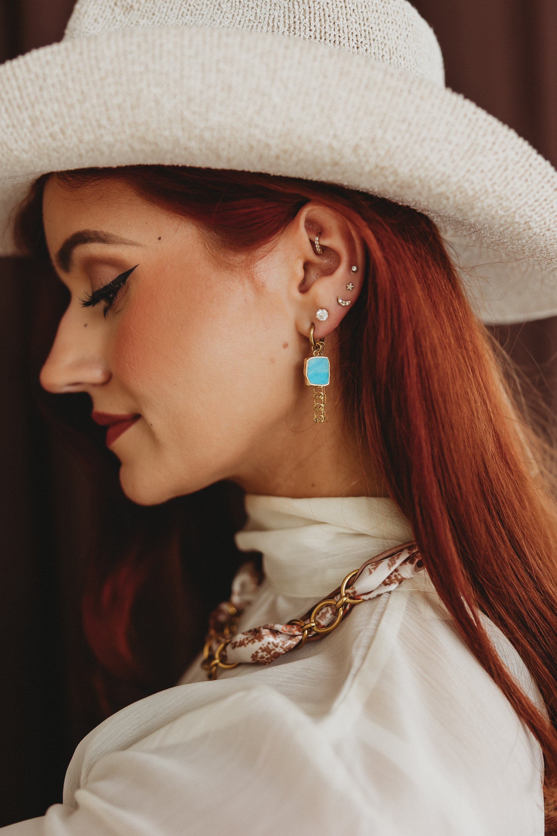 A close-up side profile of a woman with red hair wearing a white woven hat, white blouse, and a gold-toned chain necklace with a turquoise blue rectangular pendant earring.