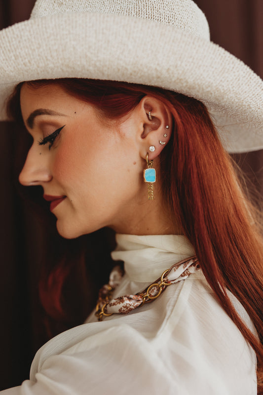 A close-up side profile of a woman with red hair wearing a white woven hat, white blouse, and a gold-toned chain necklace with a turquoise blue rectangular pendant earring.
