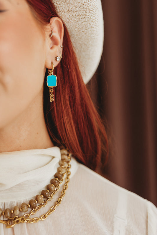 A close-up lifestyle shot of a woman wearing a gold dangle earring with a bright turquoise blue square stone, styled with a white hat, white top, and layered gold necklaces.