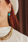 A close-up lifestyle shot of a woman wearing a gold dangle earring with a bright turquoise blue square stone, styled with a white hat, white top, and layered gold necklaces.