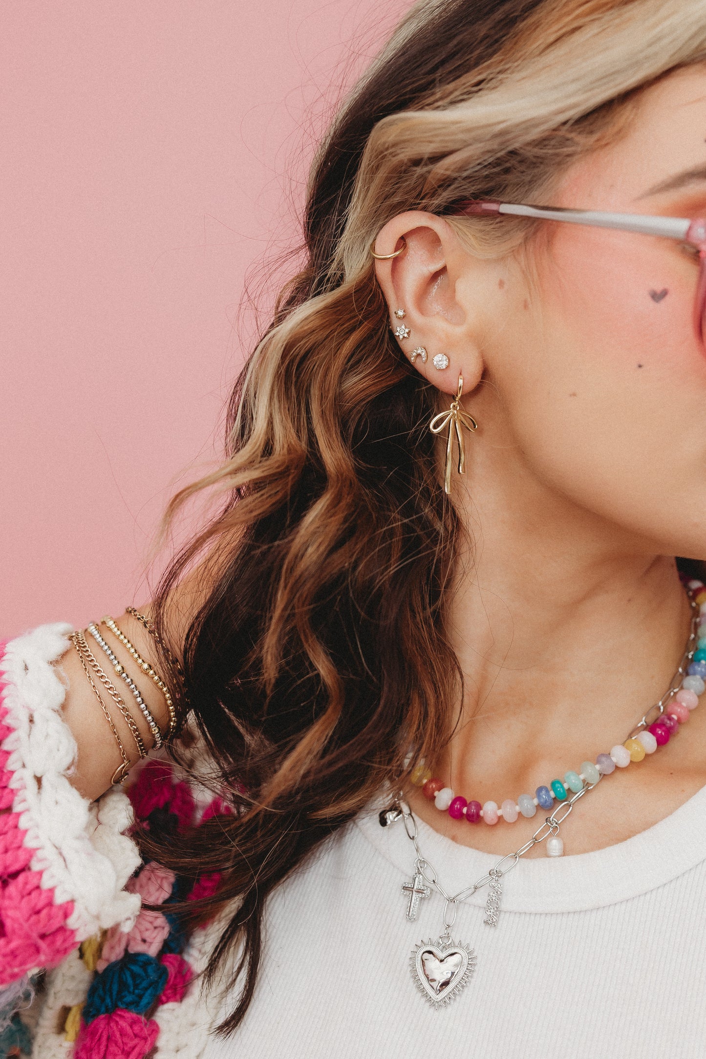Close-up of a person wearing multiple necklaces and earrings with a pink background