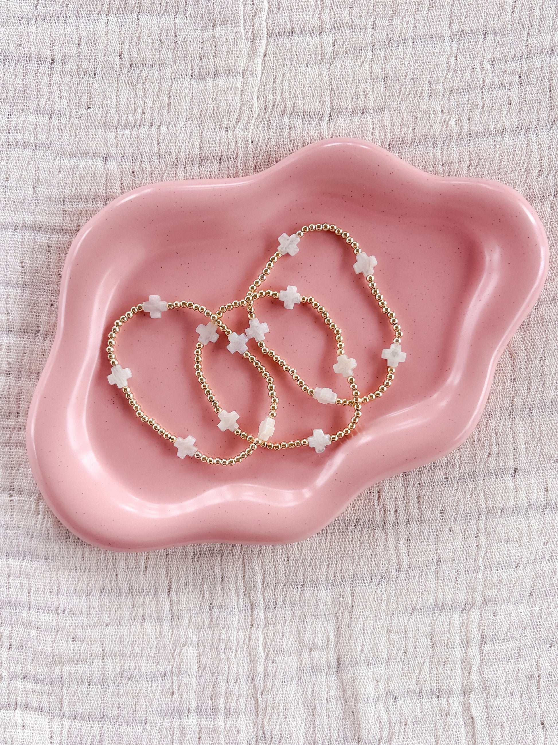 Pink ceramic dish with gold beaded white moonstone cross bracelets on a textured beige background