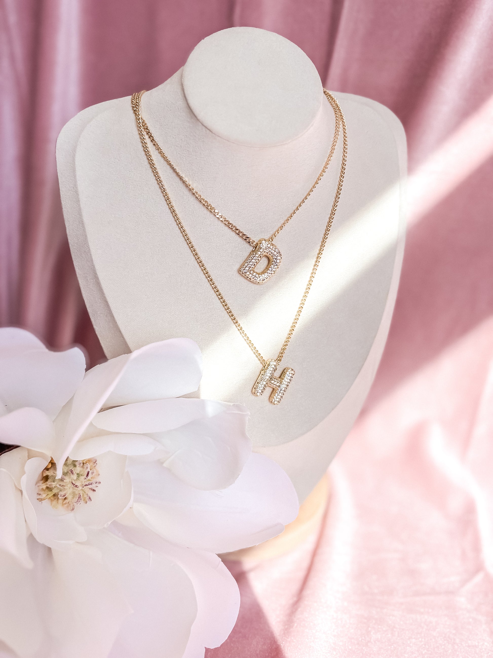 Gold necklaces with a 'D' and 'H' pendant on a mannequin bust with white flowers and pink background